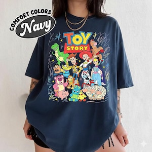 May include: Navy blue t-shirt featuring a colorful graphic of Toy Story characters. The design includes Woody, Buzz Lightyear, and other characters from the animated film. The shirt has the words "TOY STORY" in a playful font and the words "COMFORT COLORS Navy" in a circle.