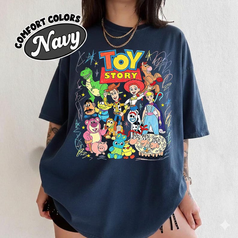 May include: Navy blue t-shirt featuring a colorful graphic of Toy Story characters. The design includes Woody, Buzz Lightyear, and other characters from the animated film. The shirt has the words "TOY STORY" in a playful font and the words "COMFORT COLORS Navy" in a circle.
