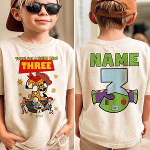 May include: A beige t-shirt featuring a colorful graphic design from the movie Toy Story. The front of the shirt says "YOU'VE GOT A FRIEND WHOS THREE" with a number 3 designed as Woody. The back of the shirt has the word "NAME" and a number 3 designed as Buzz Lightyear.