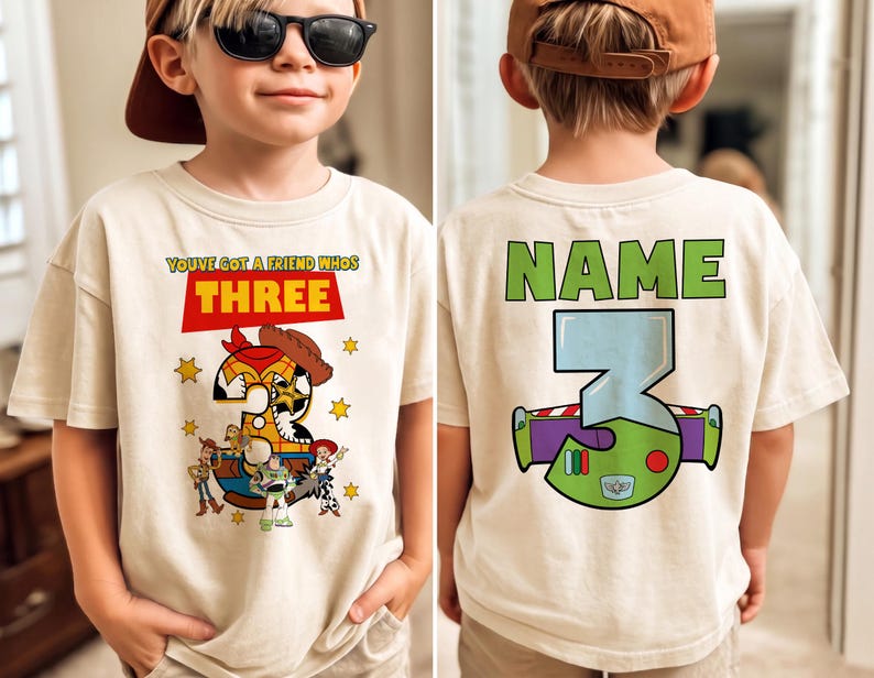 May include: A beige t-shirt featuring a colorful graphic design from the movie Toy Story. The front of the shirt says "YOU'VE GOT A FRIEND WHOS THREE" with a number 3 designed as Woody. The back of the shirt has the word "NAME" and a number 3 designed as Buzz Lightyear.