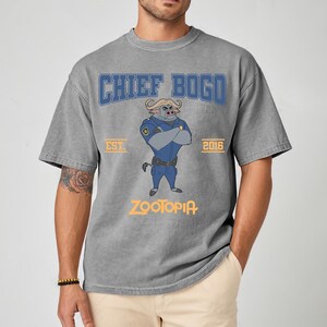 May include: Gray t-shirt featuring a cartoon illustration of Chief Bogo from Zootopia. The design includes the text "CHIEF BOGO" in blue, "EST. 2016", and "ZOOTOPIA" in orange. The shirt has short sleeves and a crew neck.
