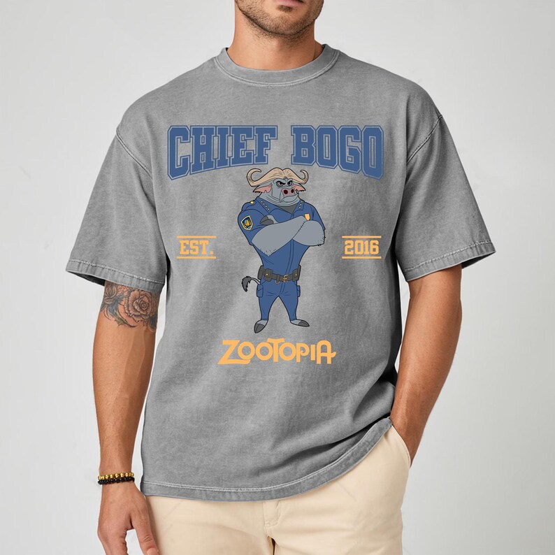 May include: Gray t-shirt featuring a cartoon illustration of Chief Bogo from Zootopia. The design includes the text "CHIEF BOGO" in blue, "EST. 2016", and "ZOOTOPIA" in orange. The shirt has short sleeves and a crew neck.