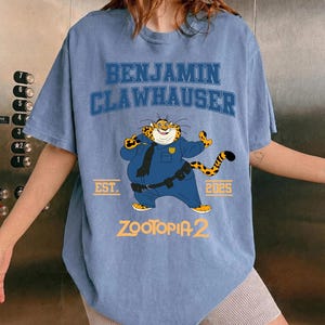 K&ouml;nnte beinhalten: Hellblaues T-Shirt mit der Aufschrift "BENJAMIN CLAWHAUSER" und einem Cartoon-Tiger in Polizeiuniform. Das Shirt hat auch den Text "EST. 2025" und "ZOOTOPIA 2" aufgedruckt.