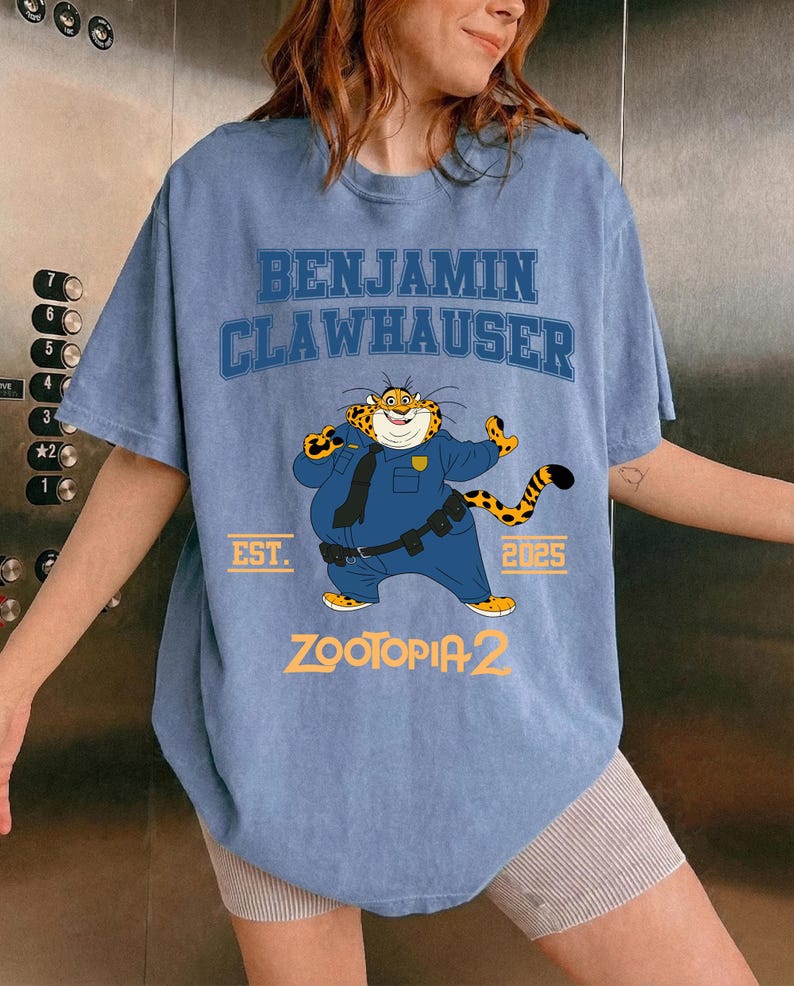 K&ouml;nnte beinhalten: Hellblaues T-Shirt mit der Aufschrift "BENJAMIN CLAWHAUSER" und einem Cartoon-Tiger in Polizeiuniform. Das Shirt hat auch den Text "EST. 2025" und "ZOOTOPIA 2" aufgedruckt.