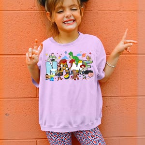 May include: A light purple sweatshirt featuring the word "NAME" in colorful block letters with cartoon character illustrations. The sweatshirt is worn by a young girl, who is smiling and giving a peace sign.