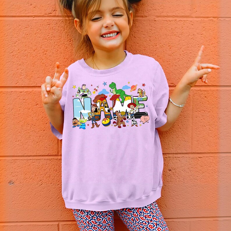 May include: A light purple sweatshirt featuring the word "NAME" in colorful block letters with cartoon character illustrations. The sweatshirt is worn by a young girl, who is smiling and giving a peace sign.