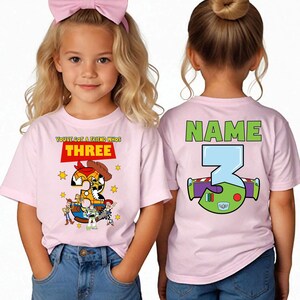 May include: Pink t-shirts with a Toy Story theme. The front of the shirt has the text "YOU'VE GOT A FRIEND WHOS THREE" with a graphic of Woody and other characters. The back has the text "NAME" and a graphic of Buzz Lightyear.
