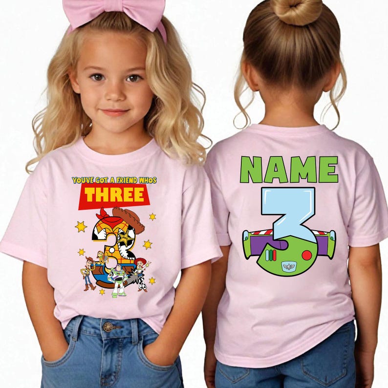 May include: Pink t-shirts with a Toy Story theme. The front of the shirt has the text "YOU'VE GOT A FRIEND WHOS THREE" with a graphic of Woody and other characters. The back has the text "NAME" and a graphic of Buzz Lightyear.