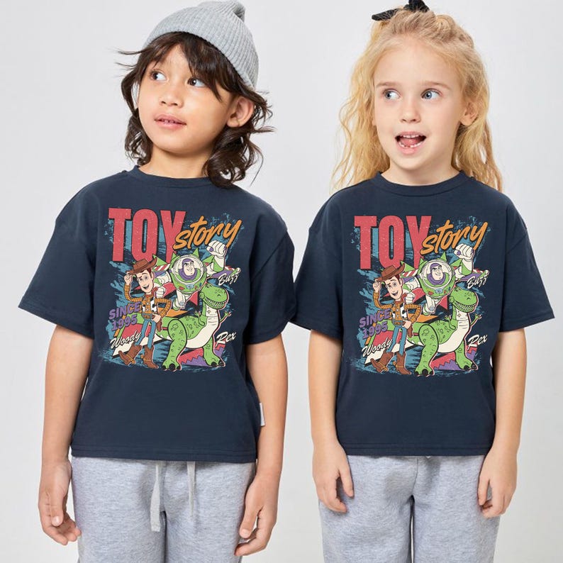 May include: Two children wearing navy blue t-shirts with a "Toy Story" graphic. The graphic features Woody, Buzz Lightyear, and Rex the dinosaur. The text "Toy Story" is in red and yellow, with the text "Since 1995" below.