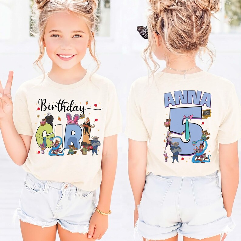 May include: A cream-colored t-shirt with the words "Birthday Girl" and the number "5" printed on it. The front of the shirt has a colorful graphic of cartoon characters. The back of the shirt has the name "Anna" printed above the number "5".