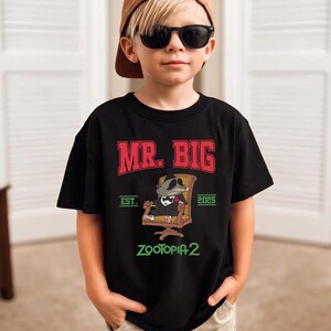 K&ouml;nnte beinhalten: Schwarzes T-Shirt mit dem Text "MR. BIG" in Rot und Wei&szlig; und "EST. 2025" darunter. Eine Zeichentrickfigur im Anzug sitzt in einem Stuhl. Das Shirt hat auch den Text "ZOOTOPIA 2" in Gr&uuml;n.