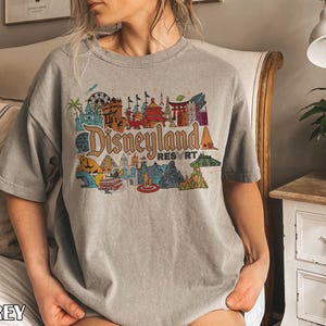 Op de afbeelding: Een grijze t-shirt met een kleurrijke Disneyland Resort afbeelding. De afbeelding toont verschillende bezienswaardigheden en attracties in een vintage stijl, met het woord "Disneyland" in een groot, gestileerd lettertype. Het woord "GREY" staat onderaan het shirt.