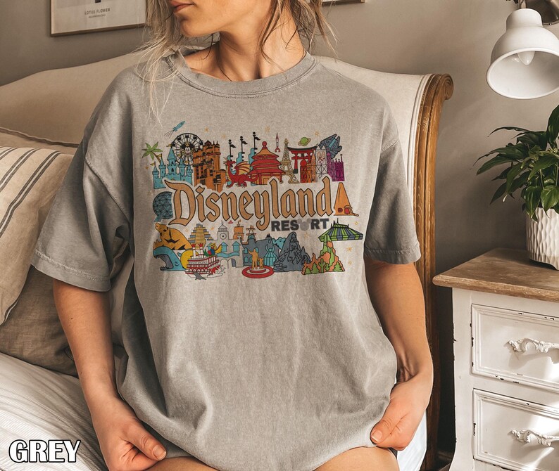 Op de afbeelding: Een grijze t-shirt met een kleurrijke Disneyland Resort afbeelding. De afbeelding toont verschillende bezienswaardigheden en attracties in een vintage stijl, met het woord "Disneyland" in een groot, gestileerd lettertype. Het woord "GREY" staat onderaan het shirt.