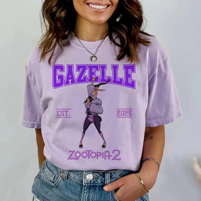 K&ouml;nnte beinhalten: Helllila T-Shirt mit dem Wort "GAZELLE" in lila Blockschrift &uuml;ber einer Gazellenillustration, die ein Mikrofon h&auml;lt. Der Text "EST. 2025" und "ZOOTOPIA 2" sind ebenfalls auf dem Shirt aufgedruckt.