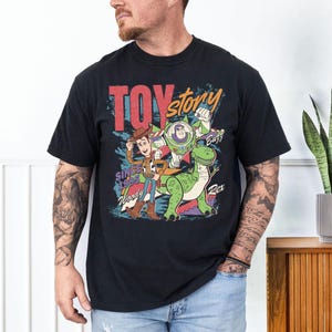 May include: Black t-shirt featuring a colorful graphic of Toy Story characters Woody, Buzz Lightyear, and Rex. The design includes the words "Toy Story" and "Since 1995". The shirt has a classic crew neck and short sleeves.