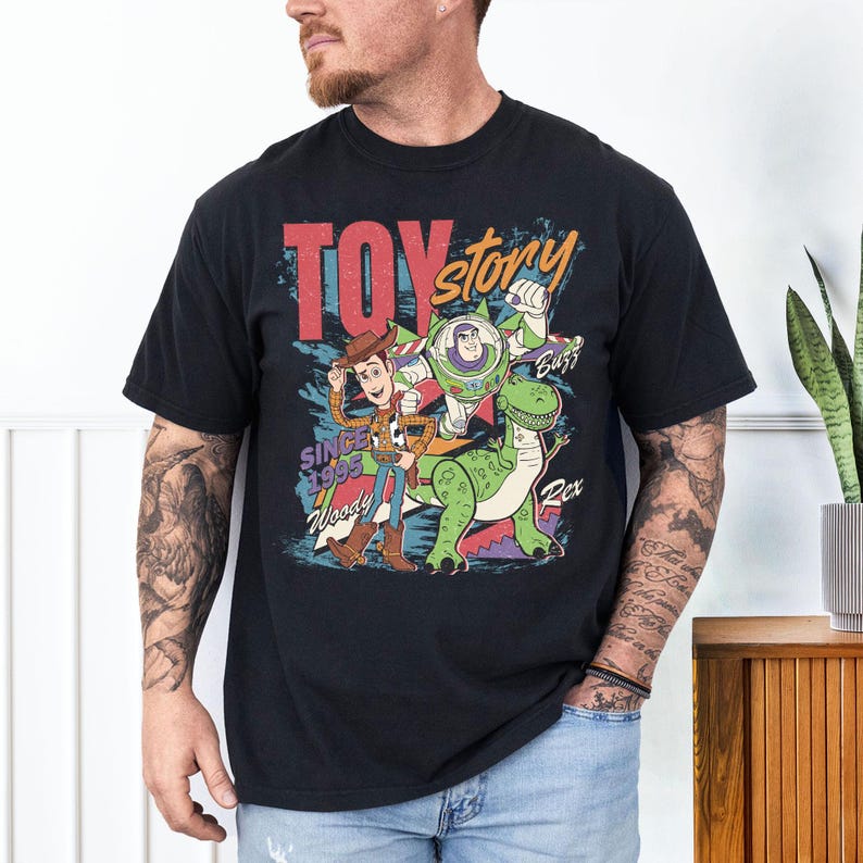 May include: Black t-shirt featuring a colorful graphic of Toy Story characters Woody, Buzz Lightyear, and Rex. The design includes the words "Toy Story" and "Since 1995". The shirt has a classic crew neck and short sleeves.