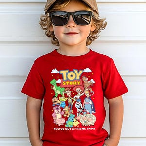 May include: A red t-shirt featuring the words "TOY STORY" in yellow and a collection of cartoon characters. Below the text is the phrase "YOU'VE GOT A FRIEND IN ME". The shirt is worn by a child wearing sunglasses and a baseball cap.