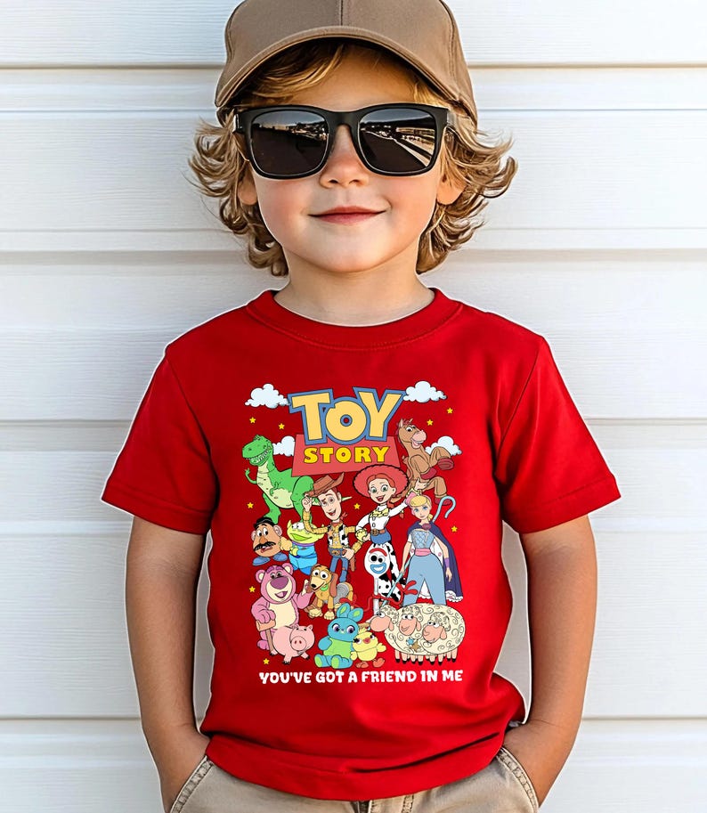 May include: A red t-shirt featuring the words "TOY STORY" in yellow and a collection of cartoon characters. Below the text is the phrase "YOU'VE GOT A FRIEND IN ME". The shirt is worn by a child wearing sunglasses and a baseball cap.