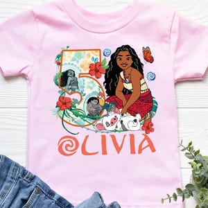 May include: Pink short-sleeved t-shirt featuring a colorful graphic of a cartoon character, flowers, and the name "OLIVIA" in orange. The number "5" is also displayed on the shirt.