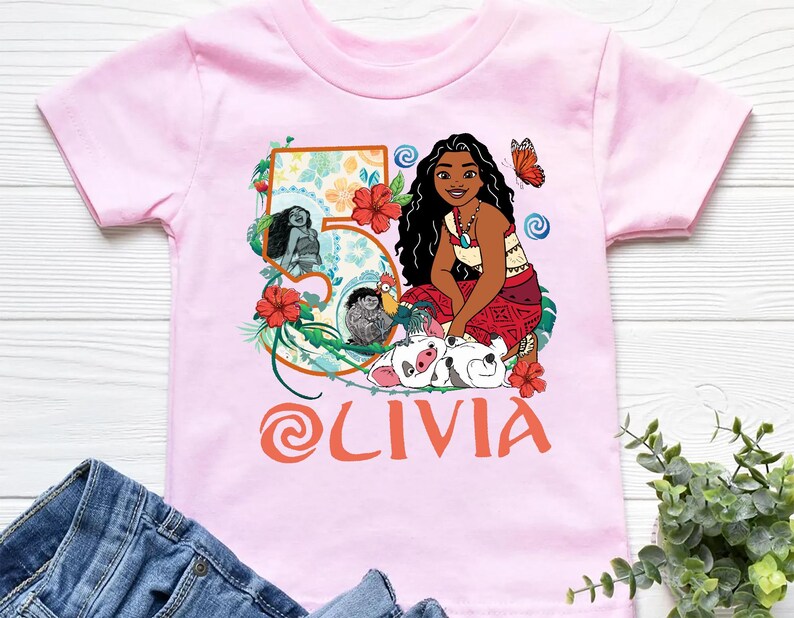 May include: Pink short-sleeved t-shirt featuring a colorful graphic of a cartoon character, flowers, and the name "OLIVIA" in orange. The number "5" is also displayed on the shirt.