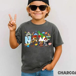 May include: Charcoal gray t-shirt featuring the word "NAME" with colorful Toy Story characters. The shirt has a vintage wash look and is made of soft material. The word "CHARCOAL" is printed at the bottom of the image.