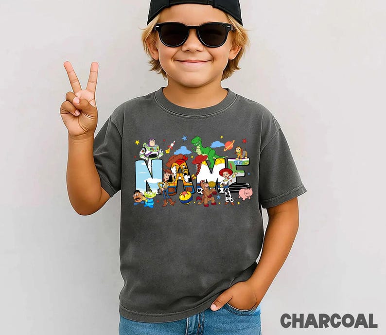 May include: Charcoal gray t-shirt featuring the word "NAME" with colorful Toy Story characters. The shirt has a vintage wash look and is made of soft material. The word "CHARCOAL" is printed at the bottom of the image.