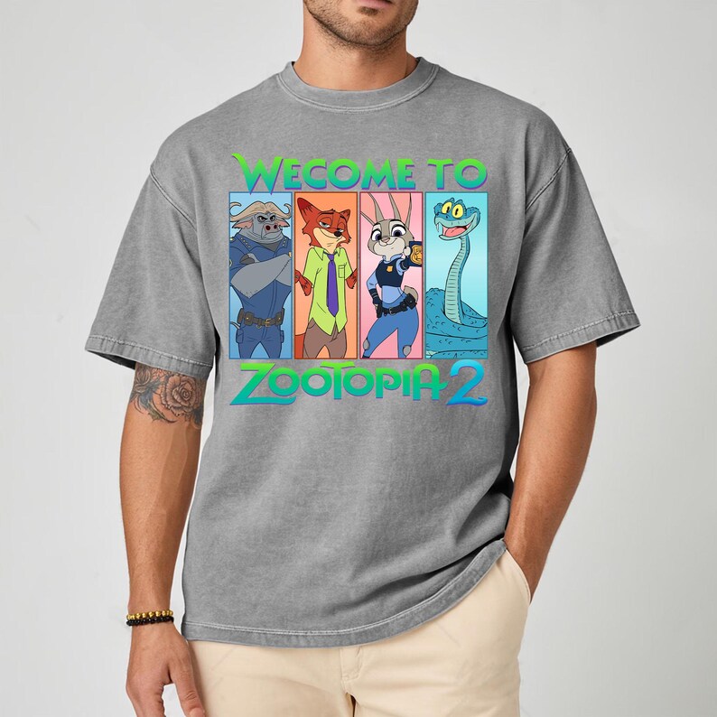 May include: Gray t-shirt featuring the words "WELCOME TO ZOOTOPIA 2" in green and blue, with cartoon characters in colorful rectangular boxes. The shirt is a casual style.