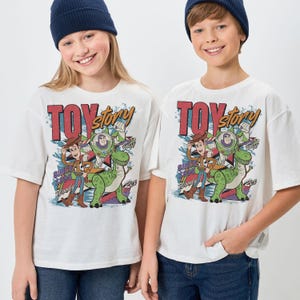 May include: Two white t-shirts with a colorful Toy Story graphic. The graphic includes the words "Toy Story" in red and gold, and images of Woody, Buzz Lightyear, and Rex the dinosaur. The shirts also have the text "Since 1995".