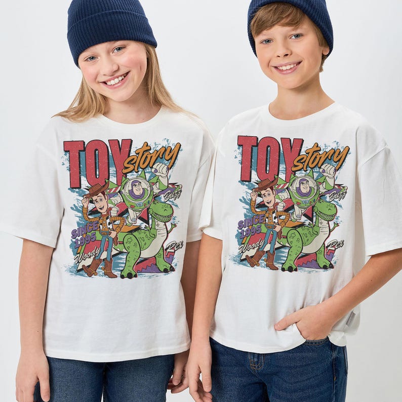 May include: Two white t-shirts with a colorful Toy Story graphic. The graphic includes the words "Toy Story" in red and gold, and images of Woody, Buzz Lightyear, and Rex the dinosaur. The shirts also have the text "Since 1995".