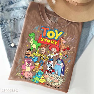 May include: Espresso-colored t-shirt featuring a colorful graphic of Toy Story characters. The design includes Woody, Buzz Lightyear, Jessie, and other characters from the animated film. The shirt is paired with blue jeans and a tan hat.