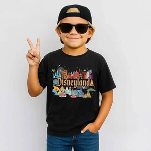 Op de afbeelding: Een zwart t-shirt met een kleurrijke Disneyland Resort-afbeelding, met iconische bezienswaardigheden en het resortlogo. Het t-shirt wordt gedragen door een persoon die een zwarte pet en zonnebril draagt.