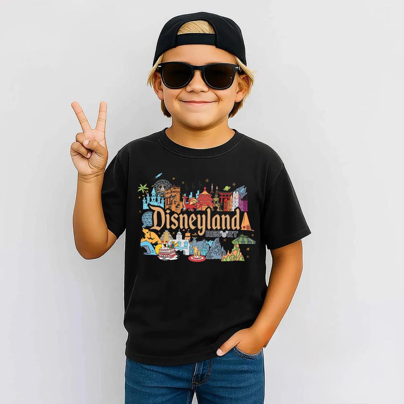Op de afbeelding: Een zwart t-shirt met een kleurrijke Disneyland Resort-afbeelding, met iconische bezienswaardigheden en het resortlogo. Het t-shirt wordt gedragen door een persoon die een zwarte pet en zonnebril draagt.