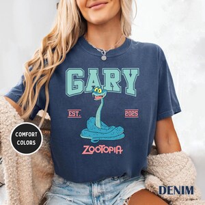 May include: Navy blue t-shirt featuring a cartoon snake named Gary from Zootopia. The design includes the name "GARY" in a teal collegiate font, the year "2025", and the word "Zootopia". The shirt has "Comfort Colors" and "Denim" text.