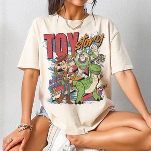 May include: Cream-colored t-shirt featuring a vintage-style graphic of Toy Story characters Woody, Buzz Lightyear, and Rex. The design includes the text "TOY STORY" in red and orange, with "SINCE 1995" and character names. A fun, nostalgic tee.