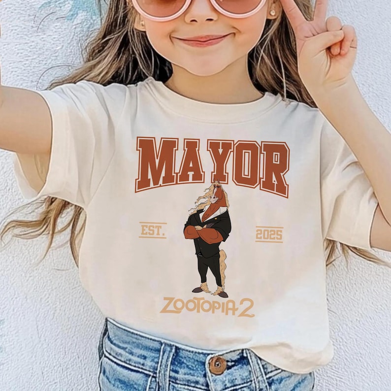 K&ouml;nnte beinhalten: Cremefarbenes T-Shirt mit dem Wort "MAYOR" in brauner Schrift im College-Stil. Darunter ein Cartoon-Pferd im Anzug mit dem Text "EST. 2025" und "ZOOTOPIA 2".