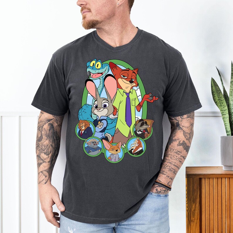 May include: Dark gray t-shirt featuring a colorful graphic of characters from the animated movie Zootopia. The design includes Judy Hopps, Nick Wilde, and other characters in a green oval frame. The shirt is a crew neck.