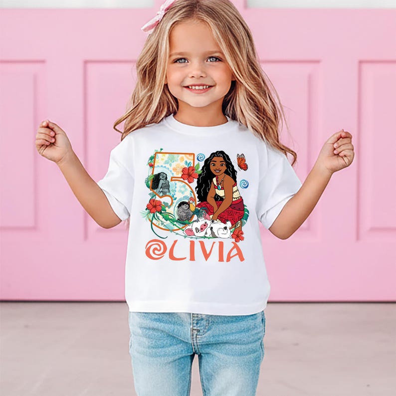 May include: White t-shirt featuring a colorful graphic of a cartoon character, flowers, and the number 5. The name "Olivia" is printed in orange script. The shirt is worn by a young girl with blonde hair and blue jeans.