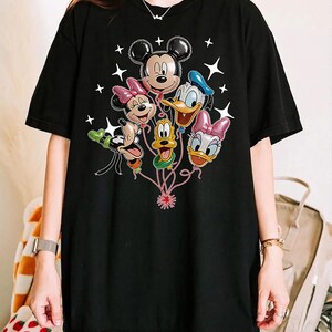 May include: Black t-shirt featuring a graphic of Mickey Mouse, Minnie Mouse, Donald Duck, Daisy Duck, Goofy, and Pluto as balloon characters. White star accents surround the balloons. The shirt is a casual, oversized style.