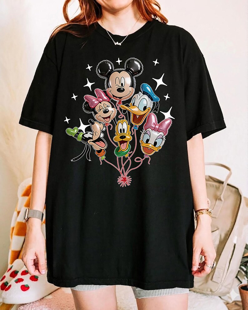 May include: Black t-shirt featuring a graphic of Mickey Mouse, Minnie Mouse, Donald Duck, Daisy Duck, Goofy, and Pluto as balloon characters. White star accents surround the balloons. The shirt is a casual, oversized style.
