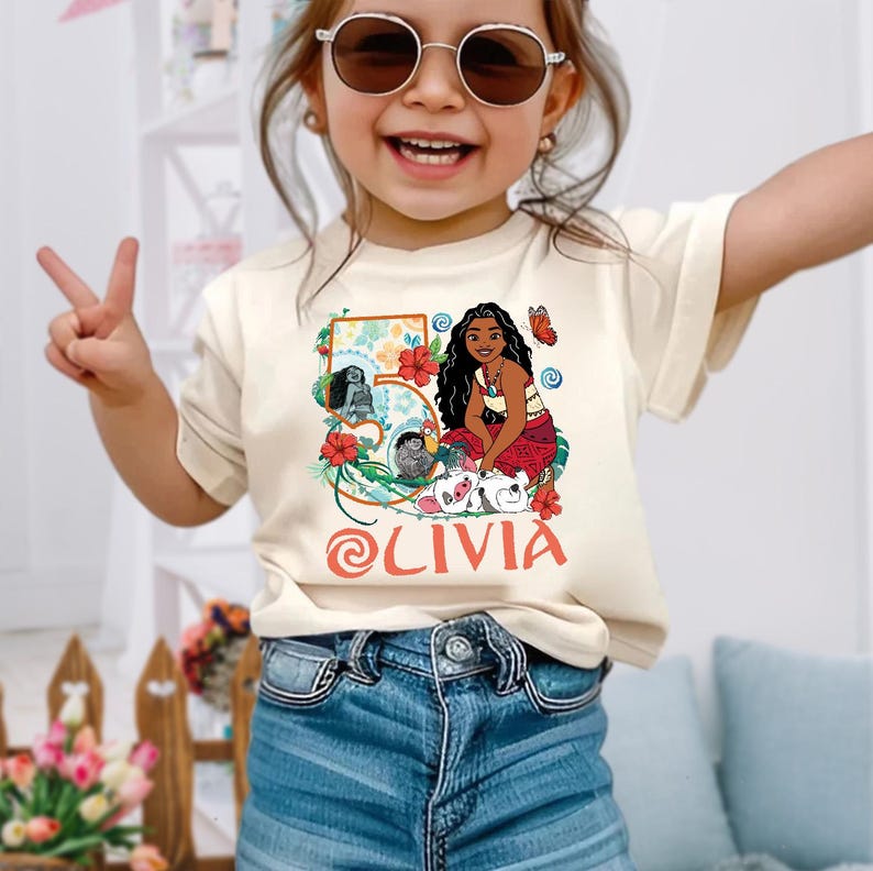 May include: A cream-colored t-shirt featuring a colorful graphic of a cartoon character with long black hair, a red skirt, and a necklace. The graphic includes the number 5, flowers, and the name "Olivia" in red. The shirt is worn by a young child.