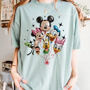 May include: A light blue t-shirt featuring a colorful graphic of Mickey Mouse, Minnie Mouse, Donald Duck, Daisy Duck, Goofy, and Pluto as balloon characters. Black star accents surround the balloons. The shirt is made of a soft material.