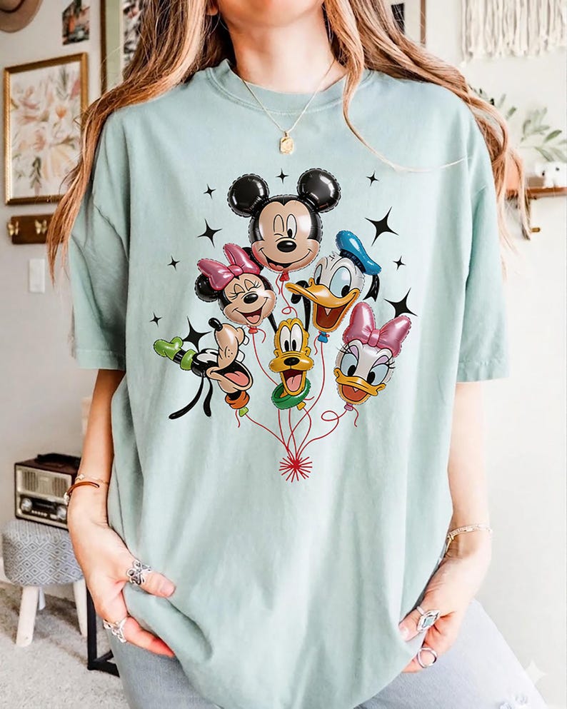May include: A light blue t-shirt featuring a colorful graphic of Mickey Mouse, Minnie Mouse, Donald Duck, Daisy Duck, Goofy, and Pluto as balloon characters. Black star accents surround the balloons. The shirt is made of a soft material.