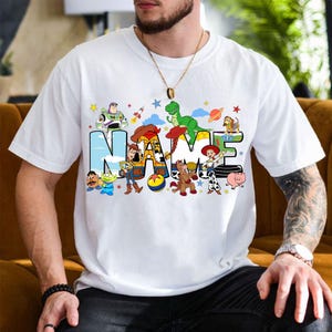 May include: White t-shirt featuring the word "NAME" in a colorful, cartoon-style design with characters from a popular animated movie. The design includes a rocket, a dinosaur, and other characters.