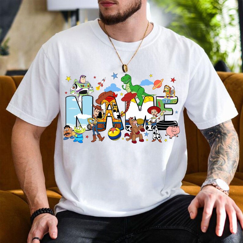 May include: White t-shirt featuring the word "NAME" in a colorful, cartoon-style design with characters from a popular animated movie. The design includes a rocket, a dinosaur, and other characters.