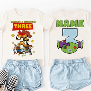 May include: Two off-white t-shirts with matching light blue denim shorts and checkered slip-on shoes. One shirt features the text "YOU'VE GOT A FRIEND WHO'S THREE" with a Toy Story graphic. The other shirt has "NAME" and a Buzz Lightyear graphic.