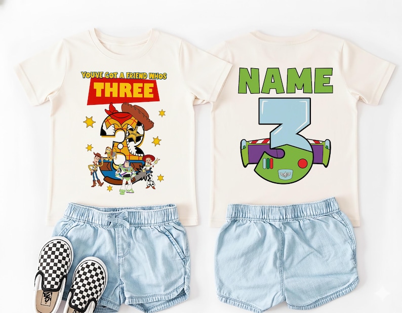 May include: Two off-white t-shirts with matching light blue denim shorts and checkered slip-on shoes. One shirt features the text "YOU'VE GOT A FRIEND WHO'S THREE" with a Toy Story graphic. The other shirt has "NAME" and a Buzz Lightyear graphic.