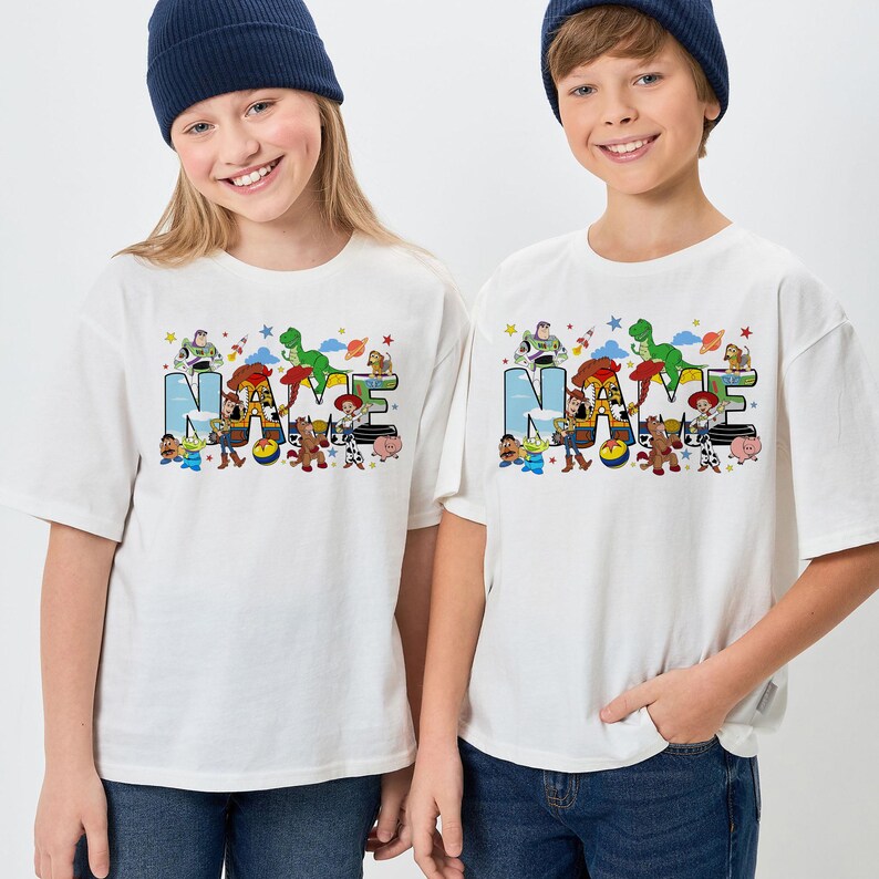May include: White t-shirts with the word "NAME" in colorful letters and cartoon characters. The shirts feature a blue, green, and red design with stars and clouds. The shirts are worn by two children.
