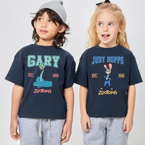 May include: Two navy blue t-shirts featuring characters from Zootopia. One shirt has "GARY" above a blue snake graphic, the other has "JUDY HOPPS" above a bunny graphic. Both shirts have "ZOOTOPIA" printed at the bottom.