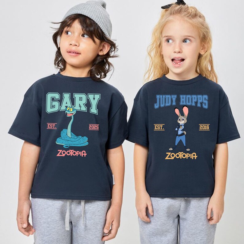 May include: Two navy blue t-shirts featuring characters from Zootopia. One shirt has "GARY" above a blue snake graphic, the other has "JUDY HOPPS" above a bunny graphic. Both shirts have "ZOOTOPIA" printed at the bottom.