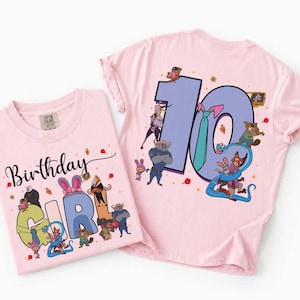 May include: Two light pink t-shirts with cartoon graphics. One shirt says "Birthday Girl" with a "2" and cartoon characters. The other shirt has a large "10" with cartoon characters. Both shirts are likely for a birthday.